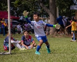 A young soccer goalkeeper in action, wearing a white and blue uniform with gloves, is preparing to intercept the ball. Two other young players, one wearing a blue and red jersey, are seated on the grass inside the goal area. The background features a lively scene with several people, trees, and a sunny day atmosphere.