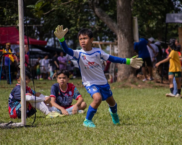 A young soccer goalkeeper in action, wearing a white and blue uniform with gloves, is preparing to intercept the ball. Two other young players, one wearing a blue and red jersey, are seated on the grass inside the goal area. The background features a lively scene with several people, trees, and a sunny day atmosphere.
