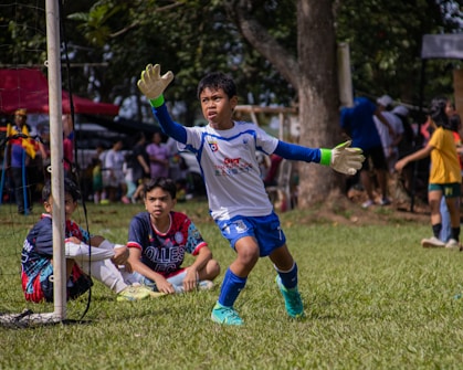 A young soccer goalkeeper in action, wearing a white and blue uniform with gloves, is preparing to intercept the ball. Two other young players, one wearing a blue and red jersey, are seated on the grass inside the goal area. The background features a lively scene with several people, trees, and a sunny day atmosphere.
