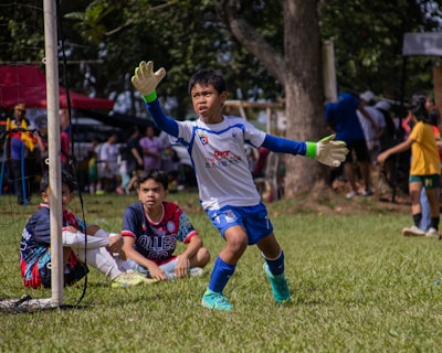 A young soccer goalkeeper in action, wearing a white and blue uniform with gloves, is preparing to intercept the ball. Two other young players, one wearing a blue and red jersey, are seated on the grass inside the goal area. The background features a lively scene with several people, trees, and a sunny day atmosphere.