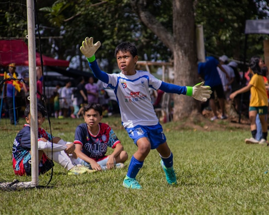 A young soccer goalkeeper in action, wearing a white and blue uniform with gloves, is preparing to intercept the ball. Two other young players, one wearing a blue and red jersey, are seated on the grass inside the goal area. The background features a lively scene with several people, trees, and a sunny day atmosphere.