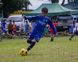 A young soccer player in blue uniform is actively kicking a yellow and black soccer ball on a grassy field. In the background, several children and adults are watching, with cars and tents nearby.