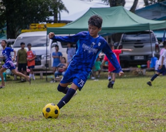 A young soccer player in blue uniform is actively kicking a yellow and black soccer ball on a grassy field. In the background, several children and adults are watching, with cars and tents nearby.