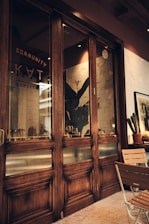 A cozy indoor setting features wooden framed glass doors with the words 'COMMUNITY' and other partially obscured text. Inside, there is a large decorative plant against a tiled wall. A wooden table and chairs are placed next to the doors. Soft lighting creates a warm atmosphere, and a framed picture and other decorative plants can be seen near the seating area.