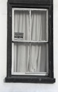 A window with dark wooden frames is set in a white wall. Behind the window, plain curtains are drawn, and a small sign hangs in the corner of the windowpane, displaying 'Furnished rooms to let'. The frame appears slightly worn, suggesting an old or vintage building.