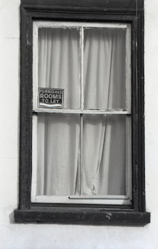 A window with dark wooden frames is set in a white wall. Behind the window, plain curtains are drawn, and a small sign hangs in the corner of the windowpane, displaying 'Furnished rooms to let'. The frame appears slightly worn, suggesting an old or vintage building.