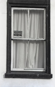 A window with dark wooden frames is set in a white wall. Behind the window, plain curtains are drawn, and a small sign hangs in the corner of the windowpane, displaying 'Furnished rooms to let'. The frame appears slightly worn, suggesting an old or vintage building.