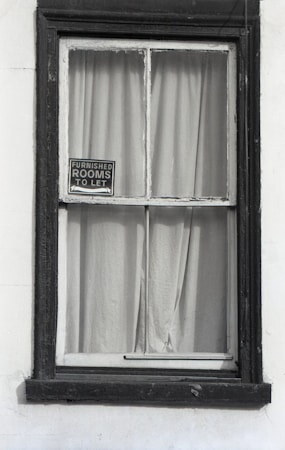 A window with dark wooden frames is set in a white wall. Behind the window, plain curtains are drawn, and a small sign hangs in the corner of the windowpane, displaying 'Furnished rooms to let'. The frame appears slightly worn, suggesting an old or vintage building.