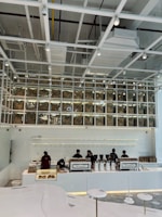 A modern coffee shop interior features a sleek, minimalist design with white and neutral tones. Several employees work behind a long counter equipped with coffee-making machines. Above the counter, transparent boxes with coffee bags labeled with a percentage sign are stacked, creating an artistic display. Overhead, the ceiling has an industrial look with exposed beams and ductwork.