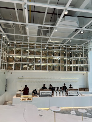 A modern coffee shop interior features a sleek, minimalist design with white and neutral tones. Several employees work behind a long counter equipped with coffee-making machines. Above the counter, transparent boxes with coffee bags labeled with a percentage sign are stacked, creating an artistic display. Overhead, the ceiling has an industrial look with exposed beams and ductwork.