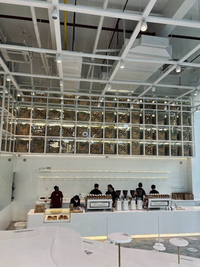A modern coffee shop interior features a sleek, minimalist design with white and neutral tones. Several employees work behind a long counter equipped with coffee-making machines. Above the counter, transparent boxes with coffee bags labeled with a percentage sign are stacked, creating an artistic display. Overhead, the ceiling has an industrial look with exposed beams and ductwork.
