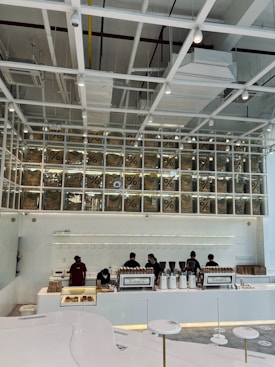 A modern coffee shop interior features a sleek, minimalist design with white and neutral tones. Several employees work behind a long counter equipped with coffee-making machines. Above the counter, transparent boxes with coffee bags labeled with a percentage sign are stacked, creating an artistic display. Overhead, the ceiling has an industrial look with exposed beams and ductwork.