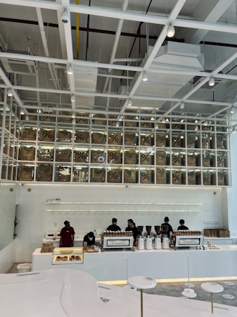 A modern coffee shop interior features a sleek, minimalist design with white and neutral tones. Several employees work behind a long counter equipped with coffee-making machines. Above the counter, transparent boxes with coffee bags labeled with a percentage sign are stacked, creating an artistic display. Overhead, the ceiling has an industrial look with exposed beams and ductwork.