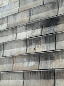 A close-up view of a textured wall made of large, rectangular stone tiles arranged in a staggered pattern. The tiles exhibit various shades of green and beige, with signs of wear and staining visible on their surfaces.