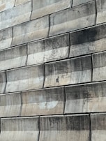 A close-up view of a textured wall made of large, rectangular stone tiles arranged in a staggered pattern. The tiles exhibit various shades of green and beige, with signs of wear and staining visible on their surfaces.