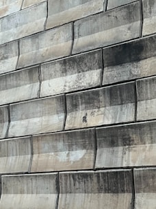 A close-up view of a textured wall made of large, rectangular stone tiles arranged in a staggered pattern. The tiles exhibit various shades of green and beige, with signs of wear and staining visible on their surfaces.