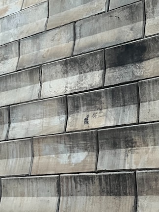 A close-up view of a textured wall made of large, rectangular stone tiles arranged in a staggered pattern. The tiles exhibit various shades of green and beige, with signs of wear and staining visible on their surfaces.