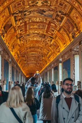 A large group of people walking through a grand hall with an ornate, illuminated ceiling featuring intricate designs and frescoes. The walls are adorned with detailed maps and artworks. One person in the center is holding up a phone to take a photo.