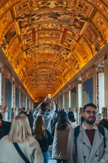 A large group of people walking through a grand hall with an ornate, illuminated ceiling featuring intricate designs and frescoes. The walls are adorned with detailed maps and artworks. One person in the center is holding up a phone to take a photo.