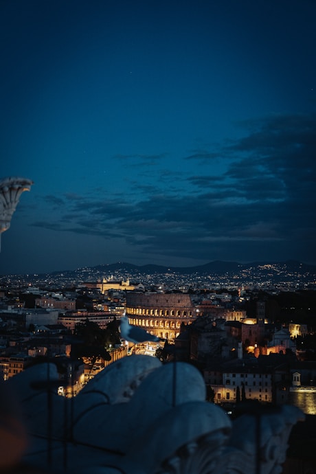 A nighttime cityscape of Rome featuring the illuminated Colosseum in the center, with city lights twinkling in the background. A large bird perched in the foreground adds depth, while the silhouette of distant hills frames the horizon under a dark blue sky.