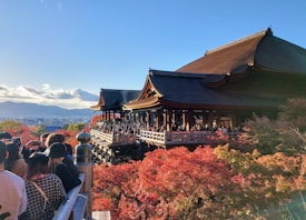 A large traditional Japanese temple structure is surrounded by vibrant autumn foliage. Visitors are gathered on the balconies, capturing the scenic beauty with their cameras. The background features a clear blue sky and distant mountains.
