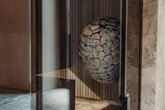 A modern interior space features a uniquely designed metal basket filled with smooth stones, mounted on a textured wall. Light wood paneling and marble surfaces create a sophisticated ambiance.