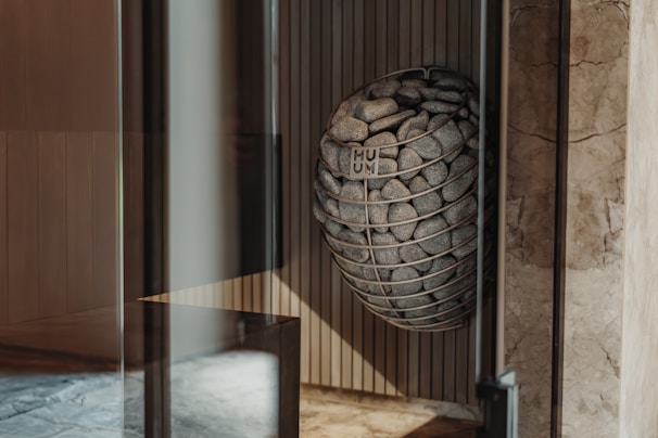 A modern interior space features a uniquely designed metal basket filled with smooth stones, mounted on a textured wall. Light wood paneling and marble surfaces create a sophisticated ambiance.