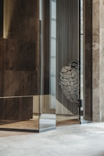 The image depicts an interior space with modern architectural elements. There are polished stone or concrete walls, with a reflective glass surface creating an intriguing layered effect. In the corner, a metallic basket holds an arrangement of stones with a label that reads 'HUUM'. The overall setting suggests a contemporary design, possibly part of a sauna or spa facility.