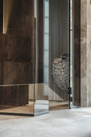 The image depicts an interior space with modern architectural elements. There are polished stone or concrete walls, with a reflective glass surface creating an intriguing layered effect. In the corner, a metallic basket holds an arrangement of stones with a label that reads 'HUUM'. The overall setting suggests a contemporary design, possibly part of a sauna or spa facility.