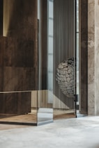 The image depicts an interior space with modern architectural elements. There are polished stone or concrete walls, with a reflective glass surface creating an intriguing layered effect. In the corner, a metallic basket holds an arrangement of stones with a label that reads 'HUUM'. The overall setting suggests a contemporary design, possibly part of a sauna or spa facility.