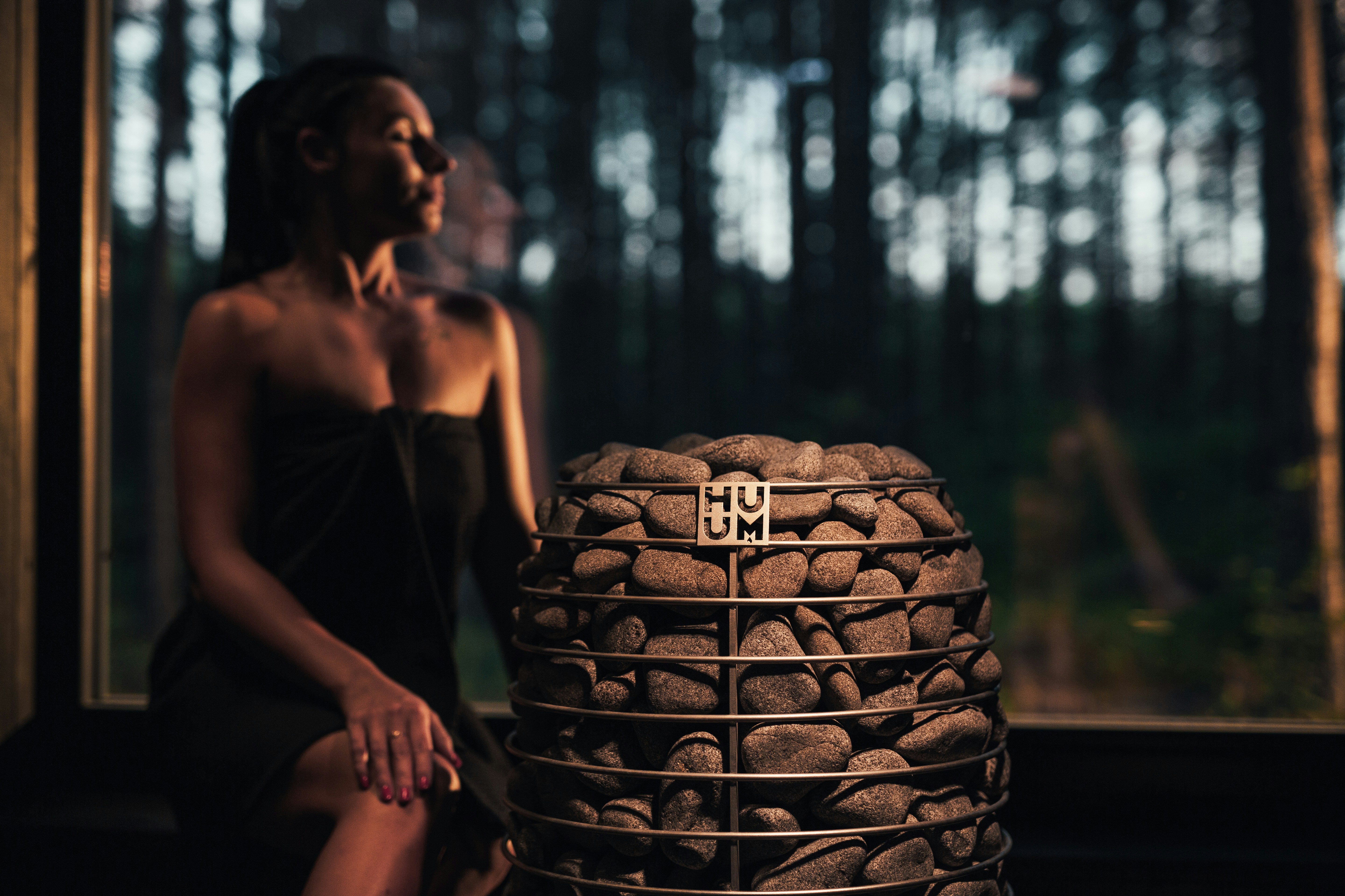 a woman sitting in front of a pile of rocks, This image highlights the HIVE Mini electric sauna heater, the perfect choice for smaller saunas without compromising on luxury. Its round, majestic design not only adds aesthetic appeal but also holds an impressive 170 kg of sauna stones. This capacity ensures a mild, long-lasting steam, making every sauna session with the HIVE Mini a memorable experience.