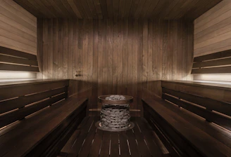 A wooden sauna room with benches lining the walls and a central heating element filled with stones. The warm tones of the wood create a cozy atmosphere, enhanced by soft lighting.