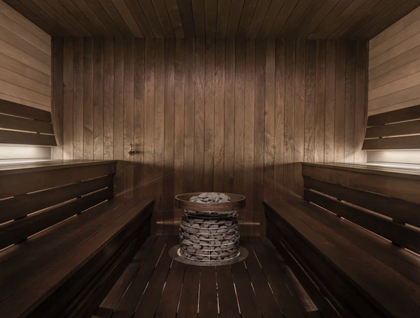 A wooden sauna room with benches lining the walls and a central heating element filled with stones. The warm tones of the wood create a cozy atmosphere, enhanced by soft lighting.