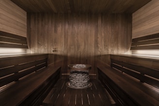 A wooden sauna room with benches lining the walls and a central heating element filled with stones. The warm tones of the wood create a cozy atmosphere, enhanced by soft lighting.