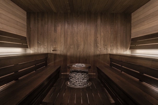A wooden sauna room with benches lining the walls and a central heating element filled with stones. The warm tones of the wood create a cozy atmosphere, enhanced by soft lighting.