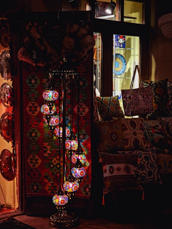 A cozy interior with a collection of colorful, intricately designed mosaic lamps hanging in a serpentine pattern. The setting includes a vibrant woven tapestry in geometric shapes, decorative plates, and patterned cushions, suggesting an eclectic, bohemian decor.