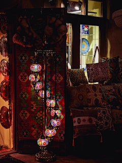 A cozy interior with a collection of colorful, intricately designed mosaic lamps hanging in a serpentine pattern. The setting includes a vibrant woven tapestry in geometric shapes, decorative plates, and patterned cushions, suggesting an eclectic, bohemian decor.