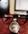 A close-up view of a wristwatch with a silver metal band placed on a desk. The wristwatch is overlaid with a pair of black eyeglasses, making the glasses appear to sit on top of the watch. In the background, a blurred computer monitor is visible.