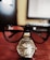 A close-up view of a wristwatch with a silver metal band placed on a desk. The wristwatch is overlaid with a pair of black eyeglasses, making the glasses appear to sit on top of the watch. In the background, a blurred computer monitor is visible.