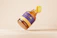 A honey bottle with a golden cap that is partially open and appears to be floating against a soft beige background. The bottle has a purple and yellow label with the words 'Honey Bottle' printed on it.