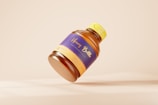 A brown plastic bottle with a bright yellow cap and a purple label, featuring the words 'Honey Bottle'. The bottle is positioned at an angle against a neutral beige background, casting a soft shadow.