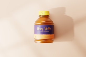 A plastic bottle containing honey with a purple and yellow label that reads 'Honey Bottle.' The bottle has a yellow cap and is placed on a light beige surface, casting a soft shadow to the right.