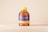 A plastic honey bottle with a yellow cap and a label featuring purple and yellow colors. The label has the words 'Honey Bottle' in a cursive font. The bottle is placed against a plain, light beige background.
