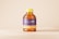 A plastic honey bottle with a yellow cap and a label featuring purple and yellow colors. The label has the words 'Honey Bottle' in a cursive font. The bottle is placed against a plain, light beige background.