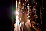 Ballet dancers in elegant tutus walk towards the stage, illuminated by bright stage lights. The scene captures the backstage area with various stage equipment visible.