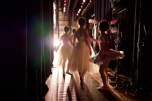 Ballet dancers in elegant tutus walk towards the stage, illuminated by bright stage lights. The scene captures the backstage area with various stage equipment visible.
