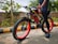 A person stands next to a black and red bicycle with thick tires, positioned on a street lined with greenery. The bicycle has prominent branding on its frame and wheels. The surroundings include small plants and trees, and the ground shows a paved surface with a yellow and black painted curb.