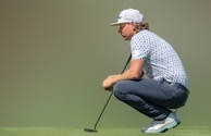 A person is crouching on a golf course holding a putter, wearing a patterned polo shirt, navy pants, and a white cap. The grass is vibrant green, and the background is blurred, creating a focus on the individual.