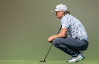 A person is crouching on a golf course holding a putter, wearing a patterned polo shirt, navy pants, and a white cap. The grass is vibrant green, and the background is blurred, creating a focus on the individual.