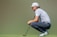A person is crouching on a golf course holding a putter, wearing a patterned polo shirt, navy pants, and a white cap. The grass is vibrant green, and the background is blurred, creating a focus on the individual.